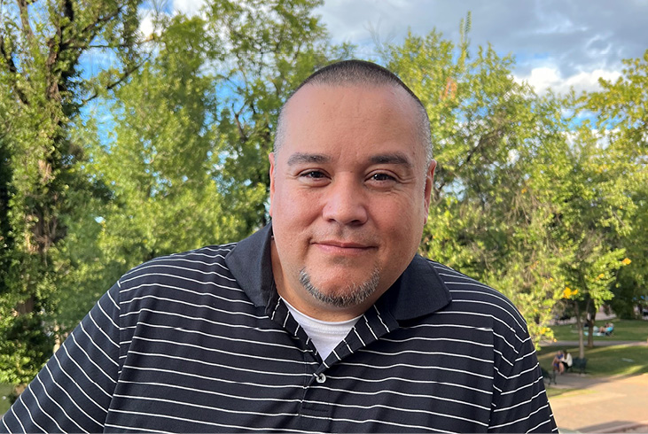 Aaron Lopez smiles for the camera. He is wearing a black and white striped shirt and is standing in front of a row of trees.